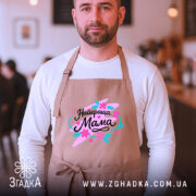 Фартух найкраща мама, бежевого кольору, на чоловікові з бородою, фон кафе, принт з написом 'Найкраща Мама' в яскравих кольорах.