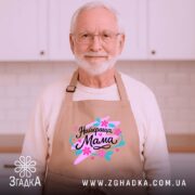 Фартух найкраща мама, бежевого кольору, зображений на дерев'яному фоні, деталі принта з написом 'Найкраща Мама' в яскравих кольорах, поряд з декором на святкову тематику.