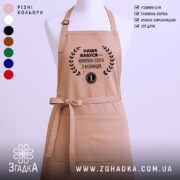 Фартух наша бабуся, бежевий колір, зображений на манекені з нейтральним фоном, принт чорного кольору з текстом і вінком.