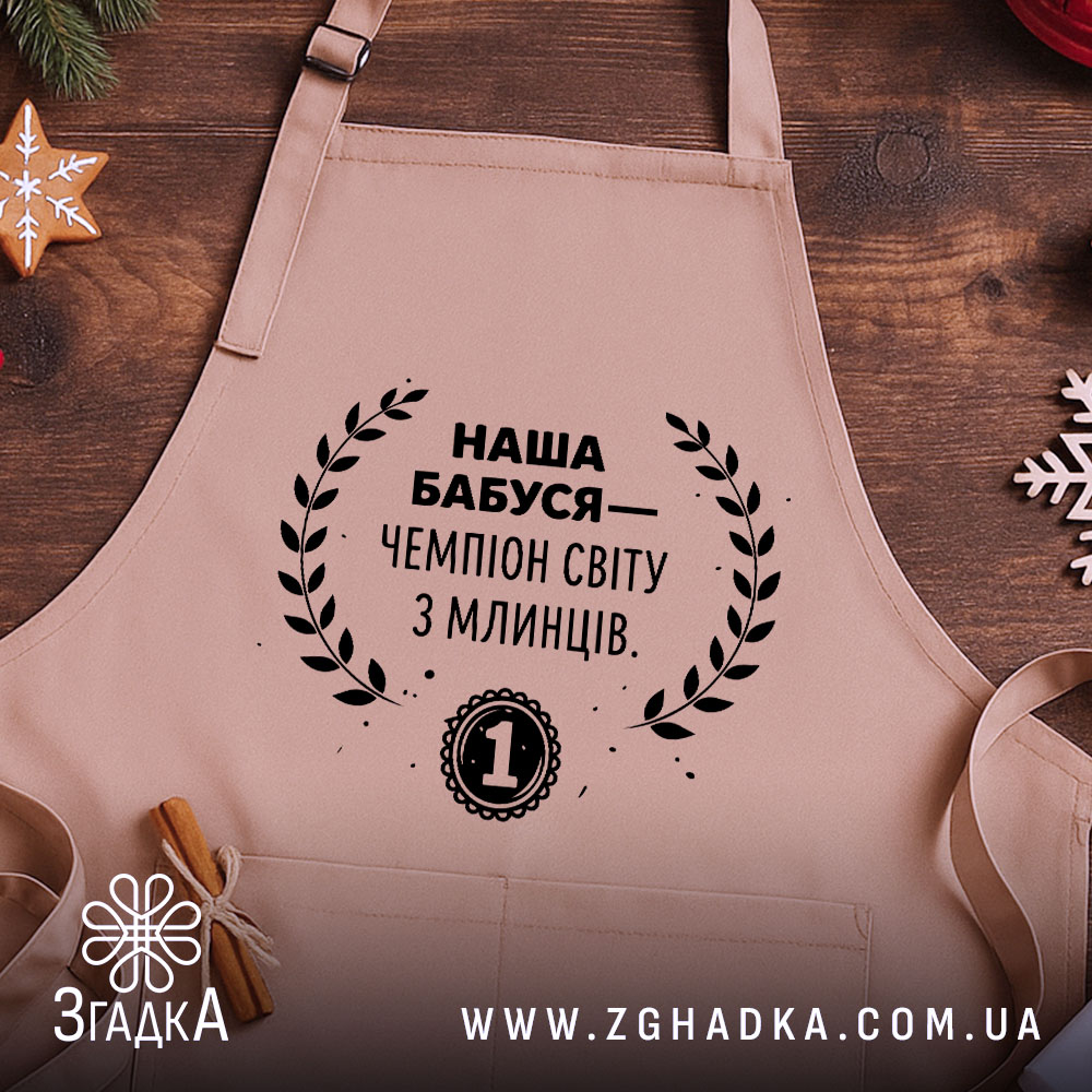 Фартух наша бабуся, бежевий колір, зображений на дерев'яному фоні з елементами декору, принт чорного кольору з текстом і вінком.