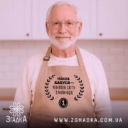 Фартух наша бабуся, бежевий колір, зображений на дерев'яному фоні з елементами декору, принт чорного кольору з текстом і вінком.