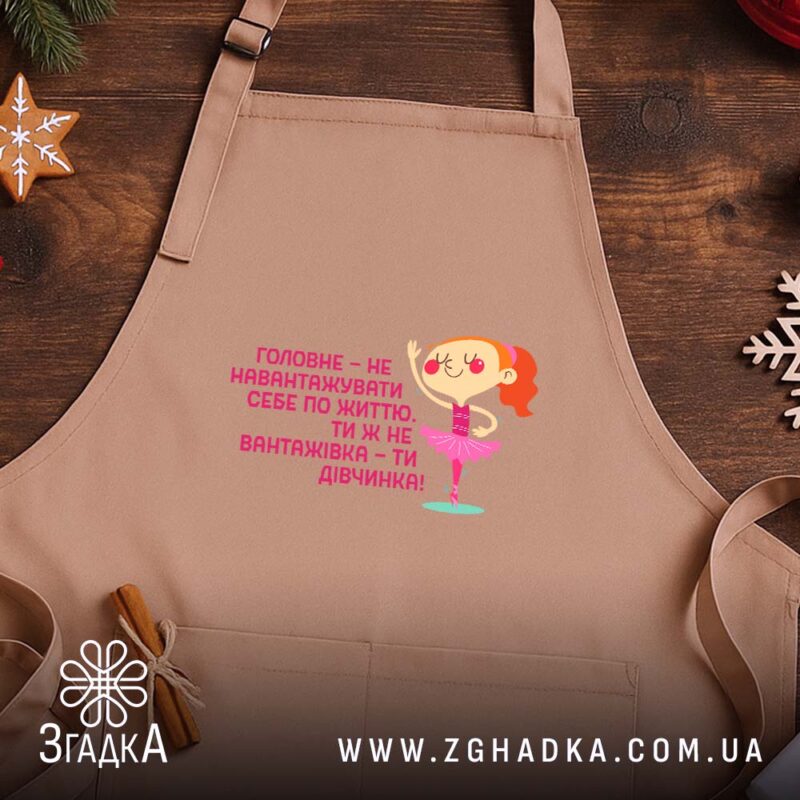 Фартух не навантажуй себе ти ж дівчинка, світло-бежевий, на дерев'яному фоні, бренд.