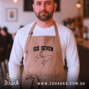 Фартух six seven жест руками, бежевий, на чоловікові, кафе на фоні, бренд.