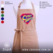 Фартух супер бабуся, бежевий колір, зображений на манекені, фон світлий, принт у формі супергеройського щита з написом 'Супер бабуся' у жовто-синіх кольорах.