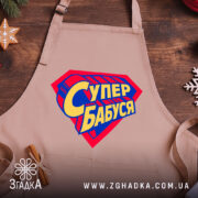 Фартух супер бабуся, бежевий колір, зображений на дерев'яному фоні, деталі принта у формі супергеройського щита з написом 'Супер бабуся' у жовто-синіх кольорах, поруч з декором у вигляді зірочки.