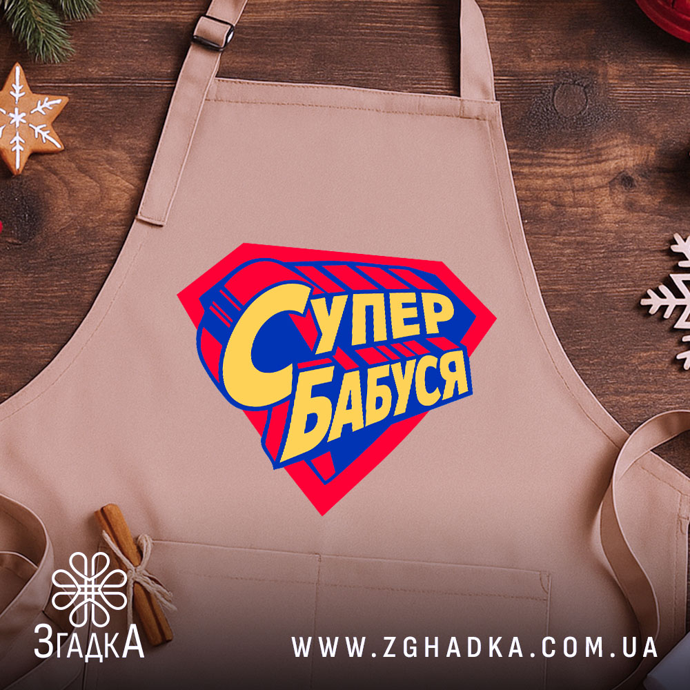 Фартух супер бабуся, бежевий колір, зображений на дерев'яному фоні, деталі принта у формі супергеройського щита з написом 'Супер бабуся' у жовто-синіх кольорах, поруч з декором у вигляді зірочки.