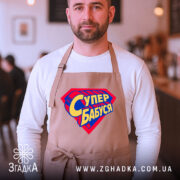 Фартух супер бабуся, бежевий колір, на чоловікові, фон кафе з розмитими людьми, принт у формі супергеройського щита з написом 'Супер бабуся' у жовто-синіх кольорах.