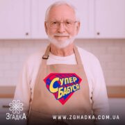 Фартух супер бабуся, бежевий колір, зображений на дерев'яному фоні, деталі принта у формі супергеройського щита з написом 'Супер бабуся' у жовто-синіх кольорах, поруч з декором у вигляді зірочки.