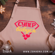 Фартух супер бабуся, світло-рожевого кольору, з верхнього ракурсу, на фоні дерев'яного столу з декором у вигляді новорічних прикрас, з принтом у вигляді червоного діаманта з написом 'СУПЕР бабуся' жовтим кольором та червоними сердечками.