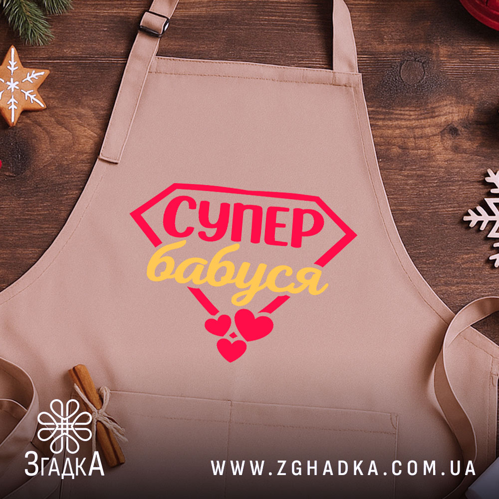 Фартух супер бабуся, світло-рожевого кольору, з верхнього ракурсу, на фоні дерев'яного столу з декором у вигляді новорічних прикрас, з принтом у вигляді червоного діаманта з написом 'СУПЕР бабуся' жовтим кольором та червоними сердечками.