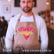 Фартух супер бабуся, бежевого кольору, з переднього ракурсу, на фоні кафе з розмитим фоном, з принтом у вигляді червоного діаманта з написом 'СУПЕР бабуся' жовтим кольором та червоними сердечками.