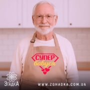 Фартух супер бабуся, світло-рожевого кольору, з верхнього ракурсу, на фоні дерев'яного столу з декором у вигляді новорічних прикрас, з принтом у вигляді червоного діаманта з написом 'СУПЕР бабуся' жовтим кольором та червоними сердечками.