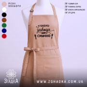 Фартух у бабусі смачно завжди. Фартух бежевого кольору, зображений з фронтального ракурсу на манекені. Фон нейтральний, без зайвих деталей. На фартуху чорний принт з написом 'У бабусі завжди смачно' та малюнками виделки і ножа.