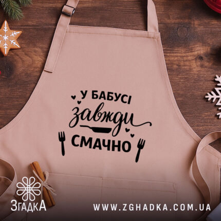 Фартух у бабусі смачно завжди. Бежевий фартух з близького ракурсу, розташований на дерев'яному фоні. На фартуху чорний принт з написом 'У бабусі завжди смачно' та малюнками кухонного приладдя. Декор на фоні включає ялинкові прикраси.