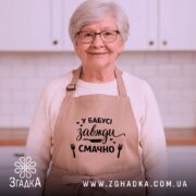 Фартух у бабусі смачно завжди. Фартух бежевого кольору, зображений з фронтального ракурсу на манекені. Фон нейтральний, без зайвих деталей. На фартуху чорний принт з написом 'У бабусі завжди смачно' та малюнками виделки і ножа.