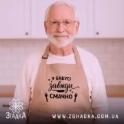 Фартух у бабусі смачно завжди. Бежевий фартух з близького ракурсу, розташований на дерев'яному фоні. На фартуху чорний принт з написом 'У бабусі завжди смачно' та малюнками кухонного приладдя. Декор на фоні включає ялинкові прикраси.