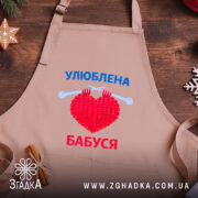 Фартух улюблена бабуся, бежевий колір, зображений на дерев'яному фоні, деталі принта з червоним серцем та синім текстом, поруч з декором у вигляді зірочки.