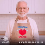 Фартух улюблена бабуся, бежевий колір, зображений на дерев'яному фоні, деталі принта з червоним серцем та синім текстом, поруч з декором у вигляді зірочки.