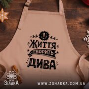 Фартух життя творить дива, бежевий, на дерев'яному фоні, бренд Згадка.