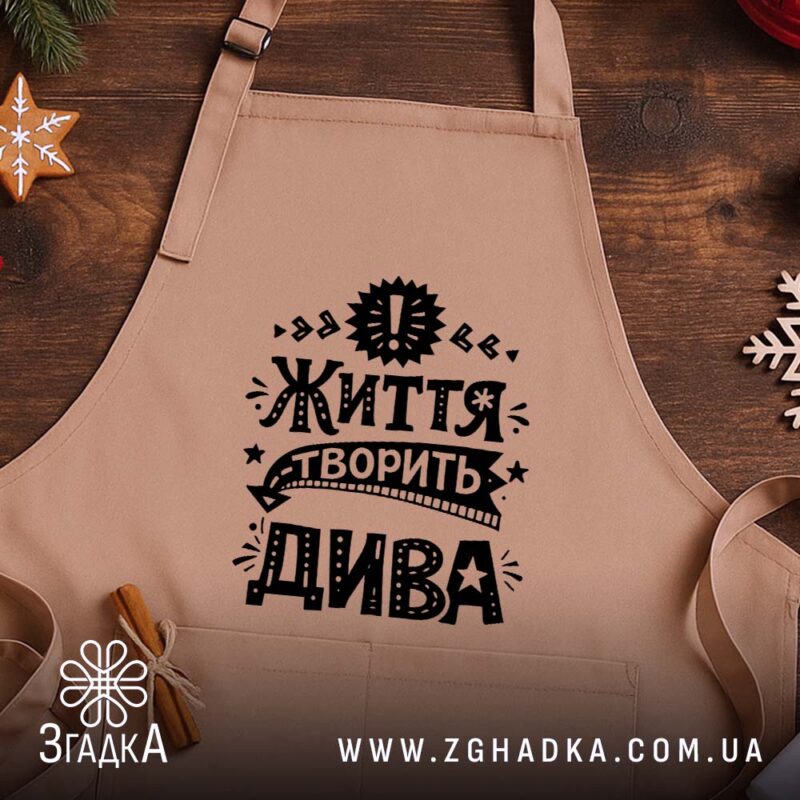 Фартух життя творить дива, бежевий, на дерев'яному фоні, бренд Згадка.