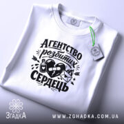 Футболка агентство розбитих сердець білого кольору, складена на поверхні, фон світло-сірий, принт чорного кольору з малюнком серця, пляшки та конверта, прикріплена етикетка з зеленим шнурком.