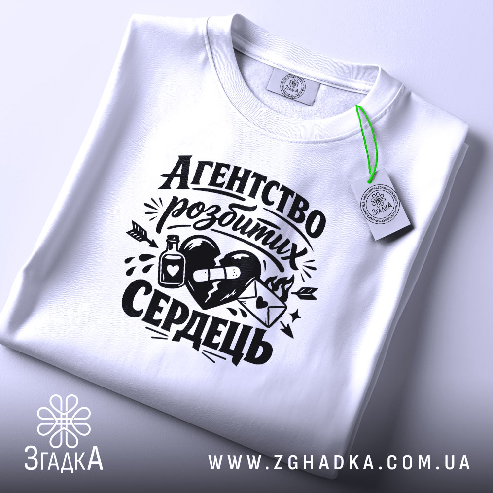 Футболка агентство розбитих сердець білого кольору, складена на поверхні, фон світло-сірий, принт чорного кольору з малюнком серця, пляшки та конверта, прикріплена етикетка з зеленим шнурком.