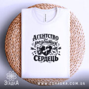 Футболка агентство розбитих сердець білого кольору, складена на плетеній кошику, фон світло-сірий, принт чорного кольору з малюнком серця, пляшки та конверта, вид зверху.