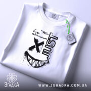 Футболка будь щасливим, біла, на плоскій поверхні, з принтом чорного кольору, напис 'just' та усмішка, тег на зеленій стрічці.