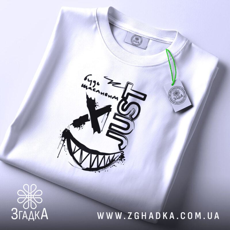 Футболка будь щасливим, біла, на плоскій поверхні, з принтом чорного кольору, напис 'just' та усмішка, тег на зеленій стрічці.