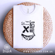 Футболка будь щасливим, біла, на плоскій поверхні, з принтом чорного кольору, напис 'just' та усмішка, розташована на плетеній основі.