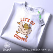 Футболка час для бешкету. Біла футболка, складена на поверхні, з принтом качки та жаби, що тримає ніж. На футболці є етикетка з зеленим шнурком. Фон світло-сірий.