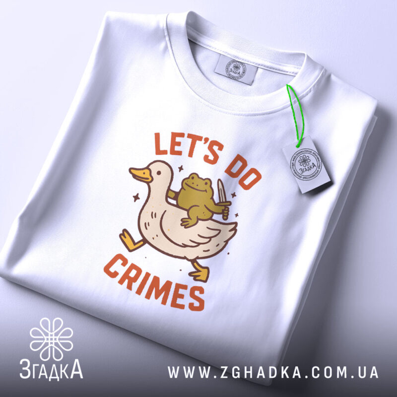 Футболка час для бешкету. Біла футболка, складена на поверхні, з принтом качки та жаби, що тримає ніж. На футболці є етикетка з зеленим шнурком. Фон світло-сірий.