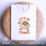 Футболка час для бешкету. Біла футболка, складена на плетеній кошику, з принтом качки та жаби, що тримає ніж. Фон світло-сірий, футболка акуратно розкладена.