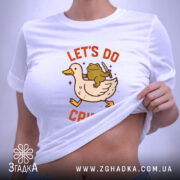 Футболка час для бешкету. Біла футболка, складена на поверхні, з принтом качки та жаби, що тримає ніж. На футболці є етикетка з зеленим шнурком. Фон світло-сірий.