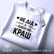 Футболка дід бабусин краш, біла, складена на столі, фон світло-сірий, принт чорний з текстом 'НЕ ДІД, а БАБУСИН КРАШ' у центрі, з етикеткою на комірці.