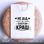 Футболка дід бабусин краш, біла, складена на плетеній кошику, фон світло-сірий, принт чорний з текстом 'НЕ ДІД, а БАБУСИН КРАШ' у центрі, з округлим комірцем.