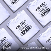 Футболка дід бабусин краш, біла, на моделі, з легким усмішкою, фон світло-фіолетовий, принт чорний з текстом 'НЕ ДІД, а БАБУСИН КРАШ' у стилі рукописного шрифту.