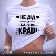 Футболка дід бабусин краш, біла, складена на столі, фон світло-сірий, принт чорний з текстом 'НЕ ДІД, а БАБУСИН КРАШ' у центрі, з етикеткою на комірці.