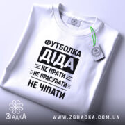 Футболка діда, біла, на столі, з коротким рукавом, фон світлий, принт чорний з текстом: 'Футболка Діда не прати не прасувати не чіпати', з етикеткою.