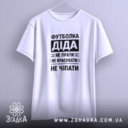 Футболка діда, біла, підвішена на вішалці, фон світлий, принт чорний з текстом: 'Футболка Діда не прати не прасувати не чіпати'.