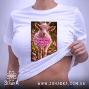 Футболка для блискучих моментів. Біла футболка, складена на поверхні, з принтом козеняти в квітах, напис 'BE THE SPARKLY ONE' яскраво виділяється. На футболці є етикетка з зеленим шнурком.