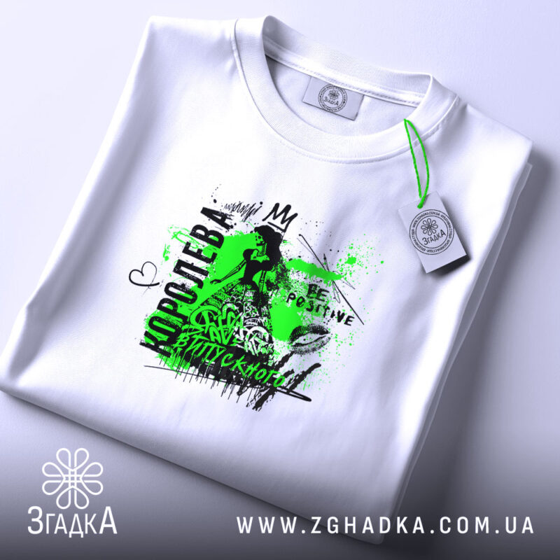 Футболка для королеви випускного, біла, на плоскій поверхні, з фоном у світло-сірому кольорі, принт з зеленими та чорними елементами, напис 'КОРОЛЕВА ВИПУСКНОГО' та графічні деталі, етикетка з зеленим шнурком.