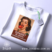 Футболка для любителів сарказму. Біла футболка, що лежить на поверхні, з принтом жінки з червоним льодяником. Фон світло-сірий, принт чіткий, текст на ньому: 'Терапія? Ні, дякую, полюбляю сарказм'.
