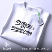 Футболка для серйозних справ. Біла футболка, складена на столі, фон світло-сірий, принт чорного кольору з текстом 'Футболка для серйозних справ' розташований по центру, на комірці є етикетка.