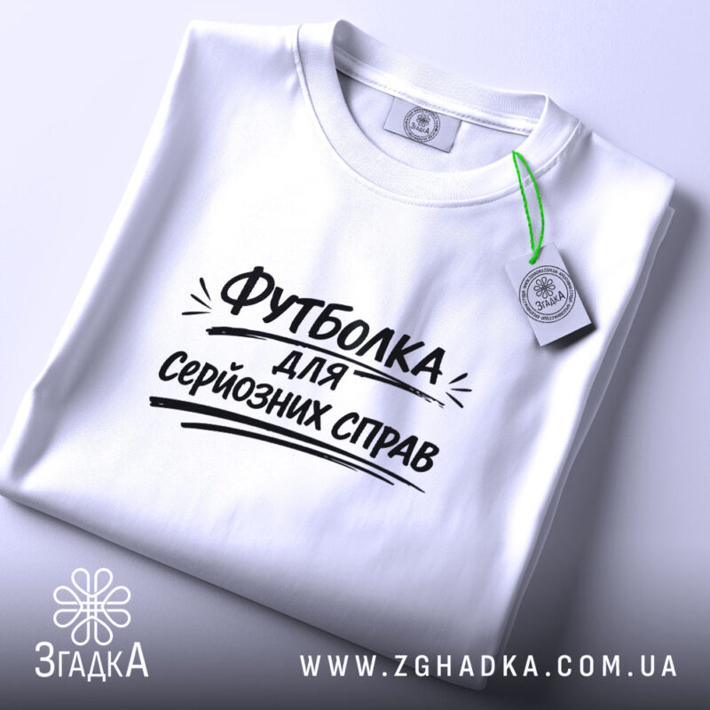 Футболка для серйозних справ. Біла футболка, складена на столі, фон світло-сірий, принт чорного кольору з текстом 'Футболка для серйозних справ' розташований по центру, на комірці є етикетка.