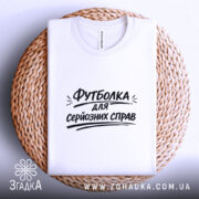 Футболка для серйозних справ. Біла футболка, складена на плетеній основі, фон білий, принт чорного кольору з текстом 'Футболка для серйозних справ' розташований по центру.