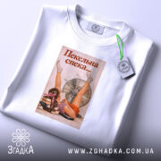 Футболка для спекотного літа, білого кольору, розкладена на поверхні, фон світлий, принт з дівчиною, що лежить біля вентилятора, з написом 'Пекельна спека...', на футболці є етикетка з зеленим шнурком.