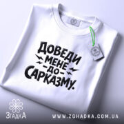 Футболка доведи мене до сарказму, біла, на столі, з етикеткою, фон світлий, принт чорного кольору з великим текстом.