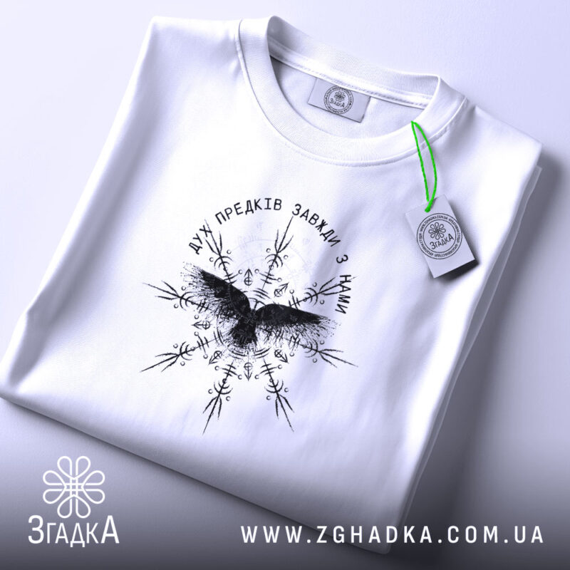 Футболка дух предків, біла, складена на столі, фон - світло-сірий, принт чорного кольору з птахом та декоративними елементами, на комірці видна етикетка.