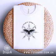 Футболка дух предків, біла, складена на плетеній кошику, фон - світло-сірий, принт чорного кольору з птахом та декоративними елементами, на комірці видна етикетка.